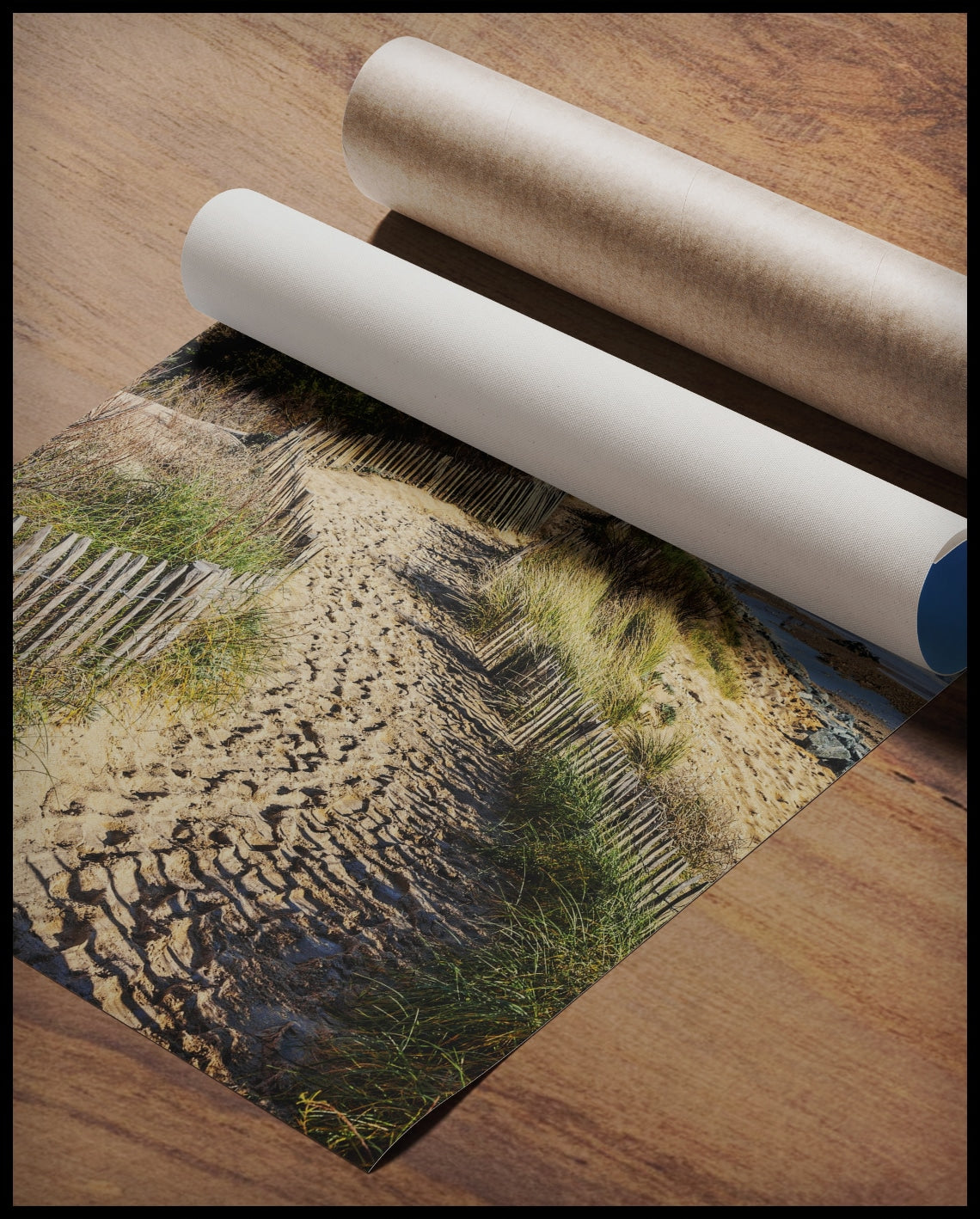 Ein Poster von einem sandigen Pfad, der sich entlang eines mit Gras und Dünen bewachsenen Küstenstreifens schlängelt, gerollt auf einer Holzoberfläche liegend