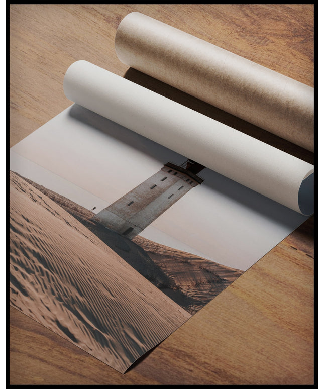 Ein Poster von einem einsamen Leuchtturm in einer sandigen Dünenlandschaft unter einem klaren Himmel, gerollt auf einer Holzoberfläche liegend