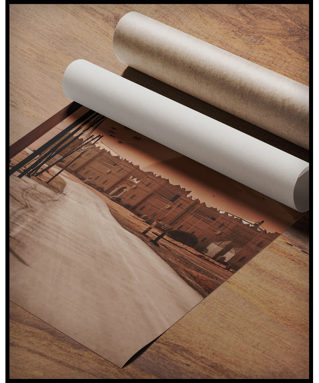 Ein Poster von einer Straße, die zu einer Siedlung am Rande einer weitläufigen Wüstenlandschaft mit hohen Sanddünen führt, gerollt auf einer Holzoberfläche liegend