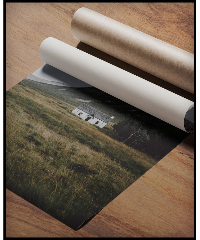 Ein Poster von einer weißen Holzhütte auf einer Wiese vor einem Berg, gerollt auf einer Holzoberfläche liegend