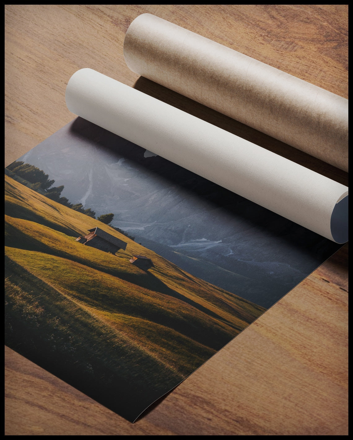 Ein Poster von zwei Holzhütten auf einer goldenen Wiese vor einer steilen, kargen Berglandschaft, gerollt auf einer Holzoberfläche liegend