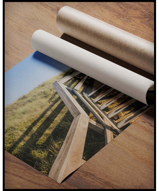 Ein Poster von einer Holztreppe die über Dünen am Strand führt, gerollt auf einer Holzoberfläche liegend