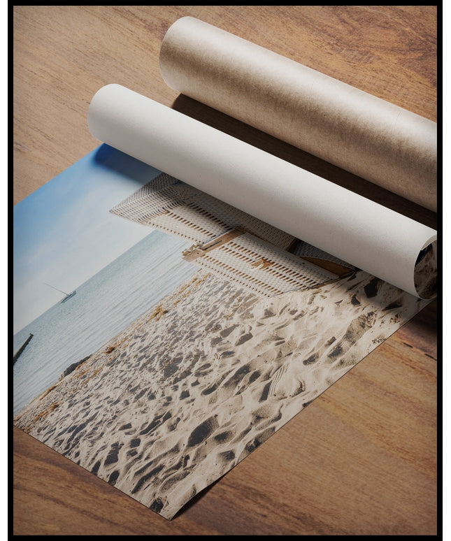 Ein Poster von einem weißen Strandkorb am Strand der in Richtung Meer gerichtet ist, gerollt auf einer Holzoberfläche liegend
