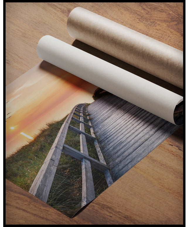 Ein Poster von einem Holzsteg im Sonnenuntergang, der über Dünen zum Strand führt, gerollt auf einer Holzoberfläche liegend