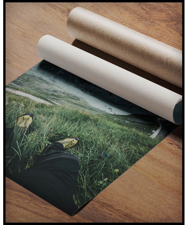 Ein Poster aus der Egoperspektive von einer Person die im Gras sitzt und auf eine Berglandschaft blickt, gerollt auf einer Holzoberfläche liegend