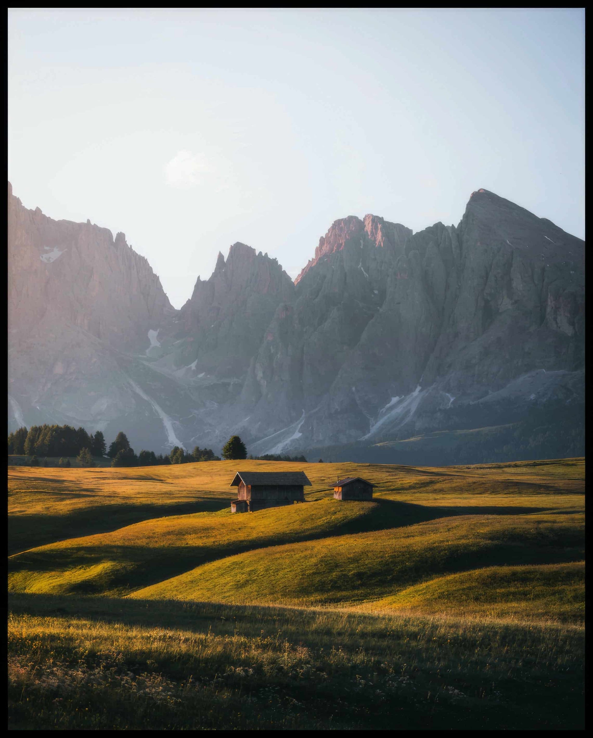 Ein Poster von zwei Holzhütten auf einer goldenen Wiese vor einer steilen, kargen Berglandschaft.