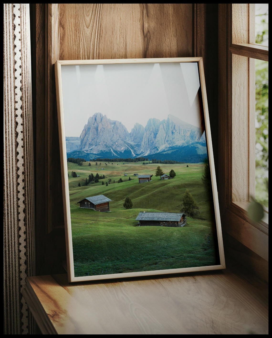 Ein gerahmtes Poster von einer grünen Alm mit kleinen Hütten und den Dolomiten im Hintergrund, vor einem Fenster an die Wand angelehnt