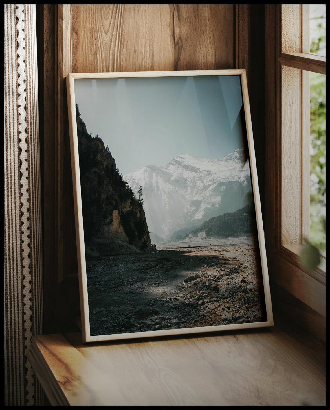 Ein gerahmtes Poster von einem breiten Flussbett in den Dolomiten, umgeben von schneebedeckten Bergen, vor einem Fenster an die Wand angelehnt