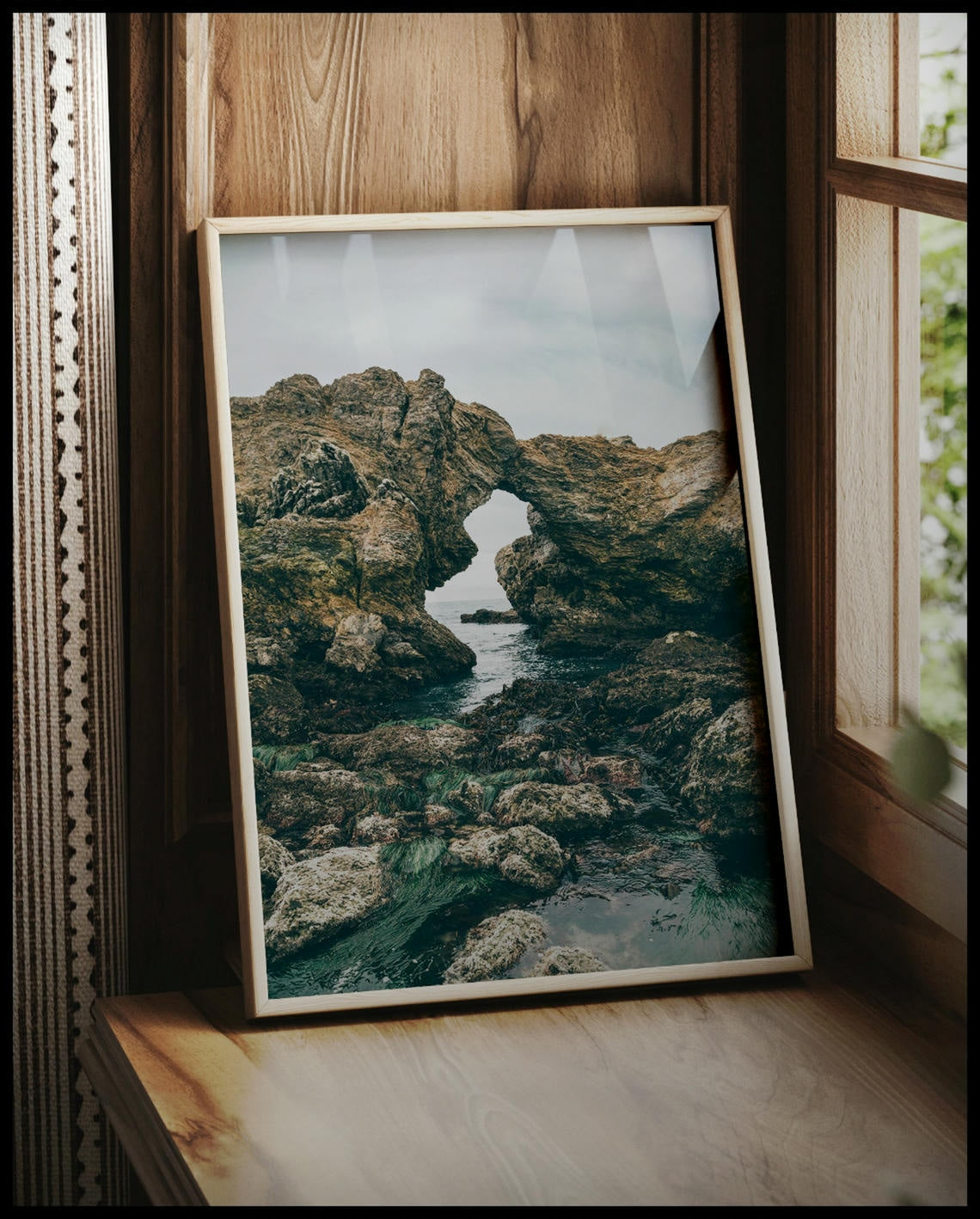 Ein gerahmtes Poster von einer felsigen Küste mit einer natürlichen Felsformation, die ein Fenster zum Meer bildet, vor einem Fenster an die Wand angelehnt