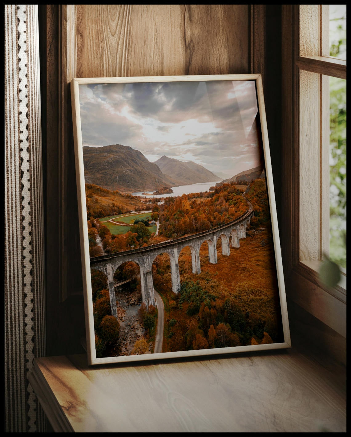 Ein gerahmtes Poster von dem Glenfinnan-Viadukt in Schottland, umgeben von herbstlicher Landschaft und Bergen im Hintergrund, vor einem Fenster an die Wand angelehnt