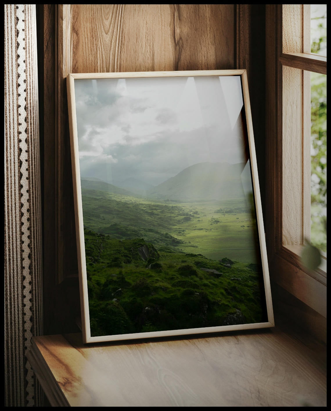 Ein gerahmtes Poster von einer grünen Landschaft in Irland mit Sonnenstrahlen, vor einem Fenster an die Wand angelehnt