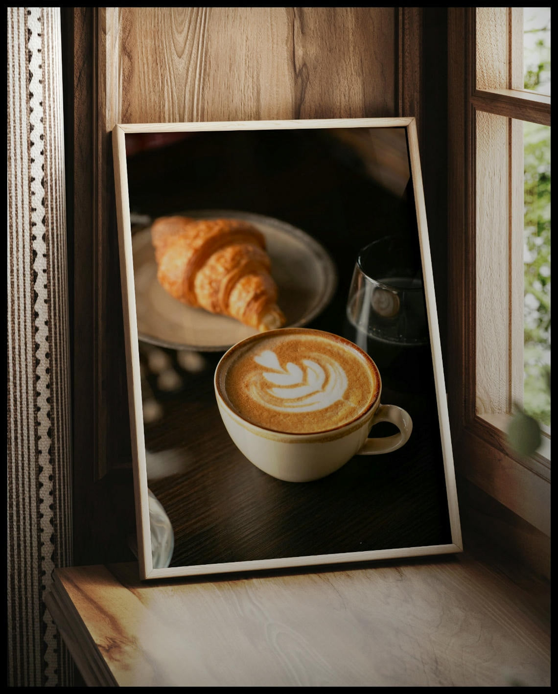 Ein gerahmtes Poster von einer Tasse Kaffee Latte neben einem Croissant und einem Glas Wasser, vor einem Fenster an die Wand angelehnt