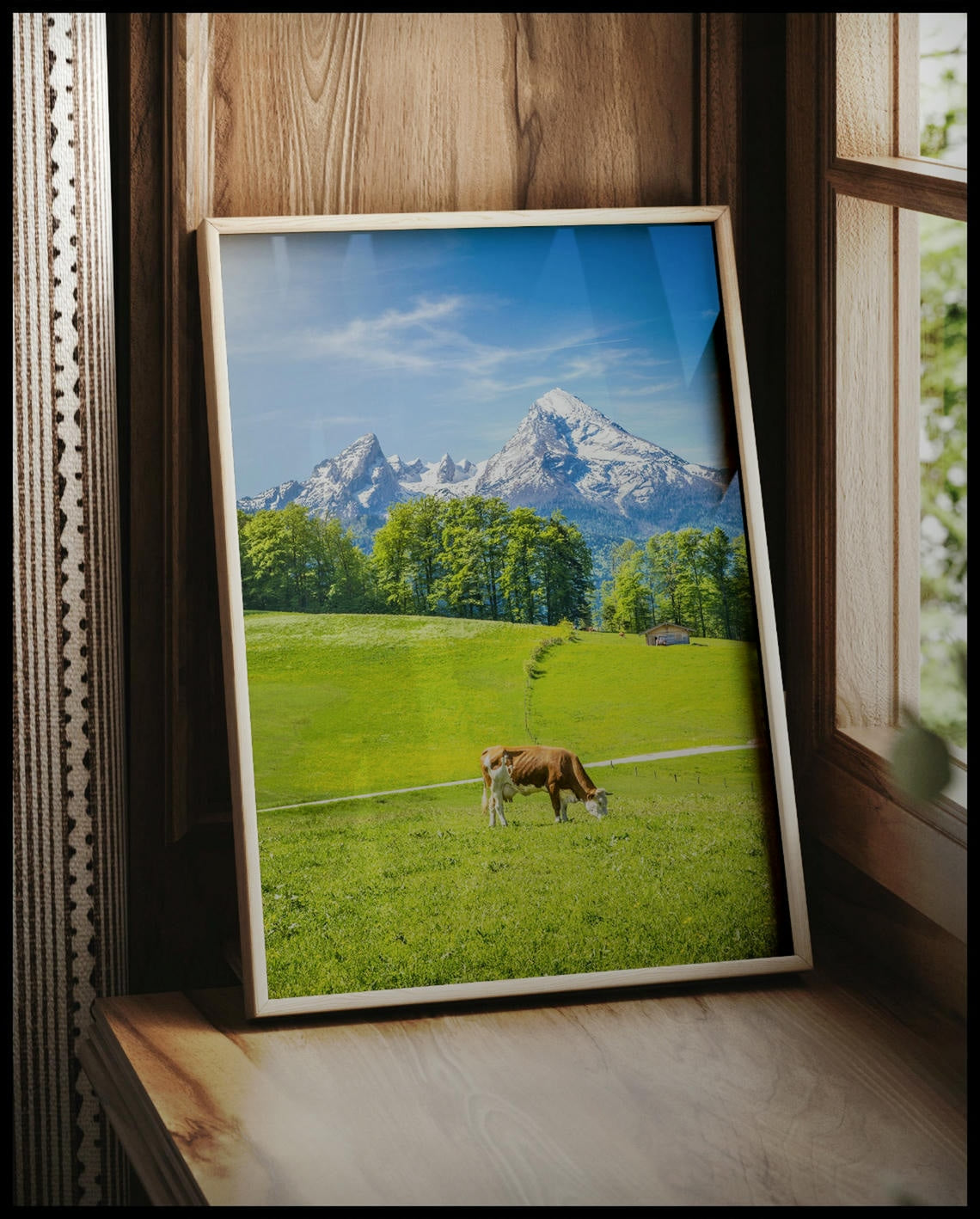 Ein gerahmtes Poster von einer Kuh auf einer grünen Wiese vor einer Alpenlandschaft, vor einem Fenster an die Wand angelehnt