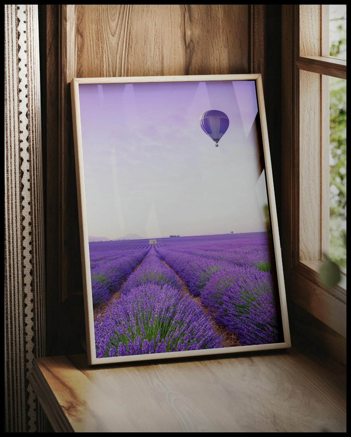 Ein gerahmtes Poster von einem Lavendelfeld und einem Heißluftballon mit lila-weißem Muster, der über dem Feld schwebt, vor einem Fenster an die Wand angelehnt