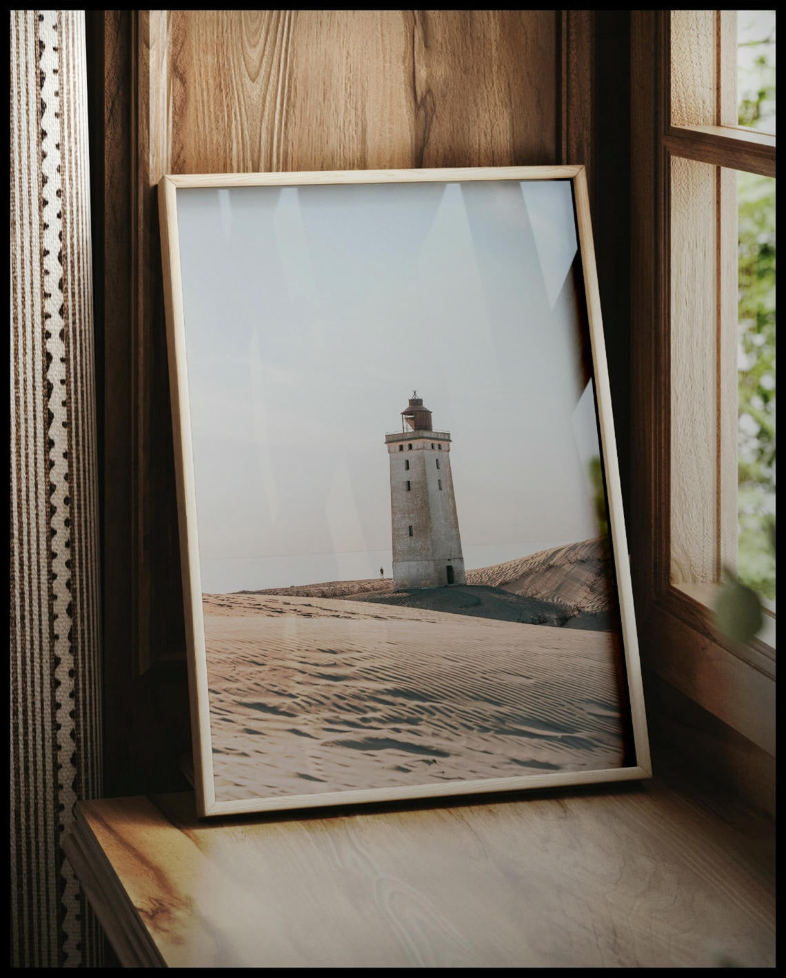 Ein gerahmtes Poster von einem einsamen Leuchtturm in einer sandigen Dünenlandschaft unter einem klaren Himmel, vor einem Fenster an die Wand angelehnt