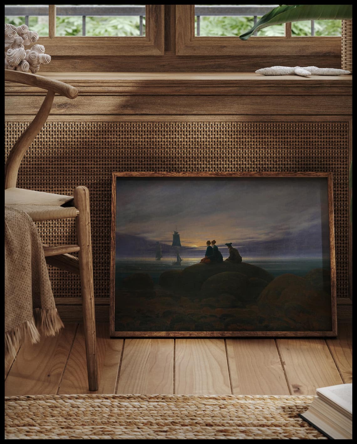 Ein gerahmter Kunstdruck von drei Personen am Meeresstrand die auf einem Felsen sitzen und den Mondaufgang betrachten, vor einem Fenster an die Wand angelehnt