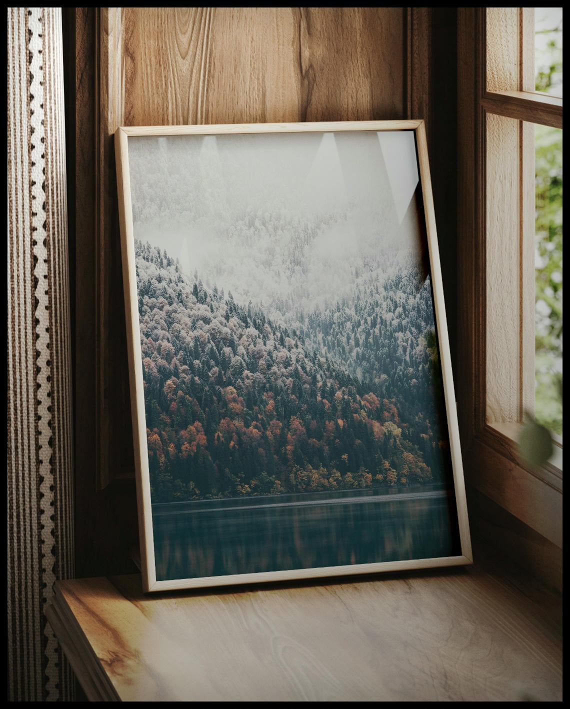 Ein gerahmtes Poster von einem nebeligen Nadelwald, der stellenweise von Schnee bedeckt ist und an einen See grenzt, vor einem Fenster an die Wand angelehnt