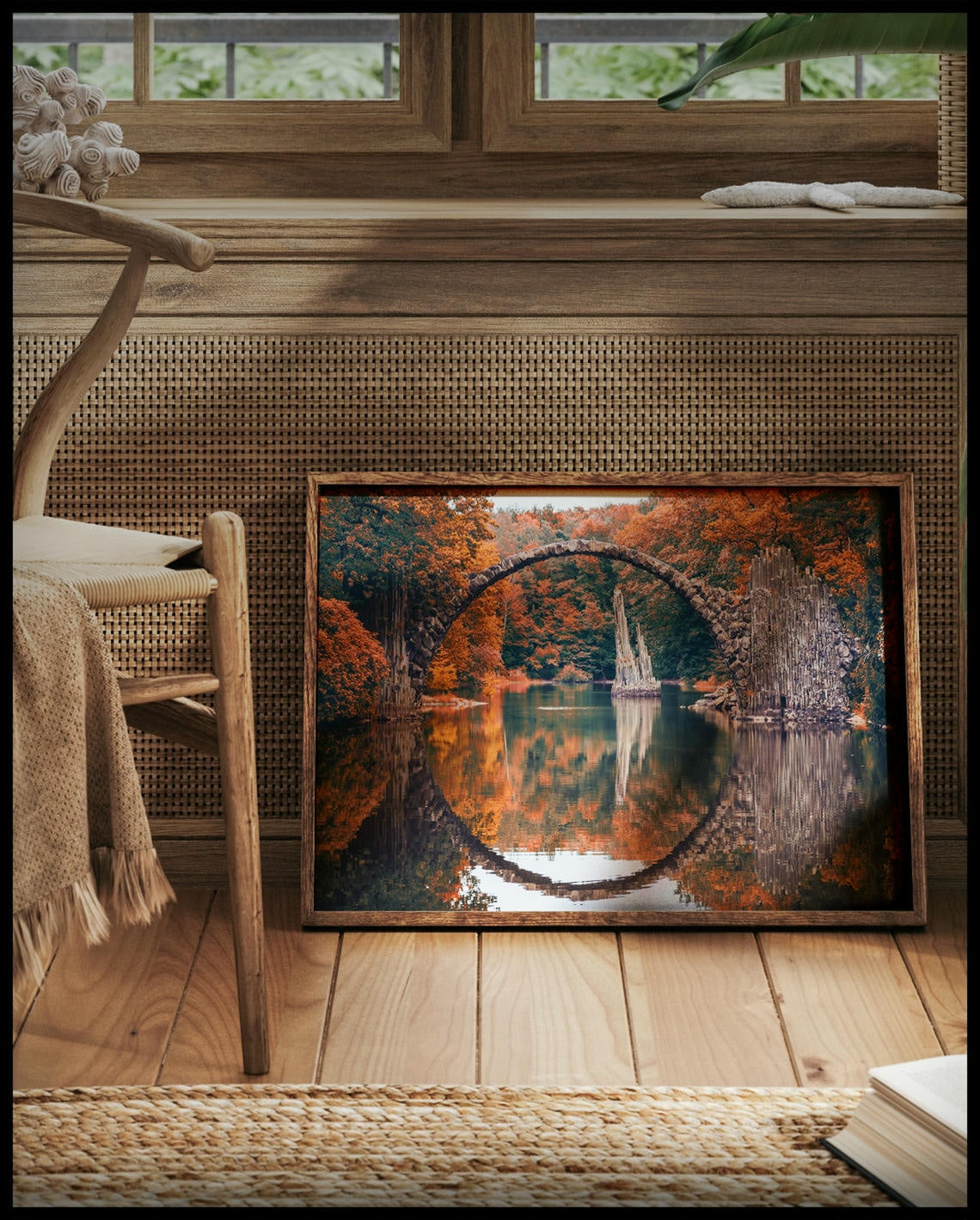 Ein gerahmtes Poster von der Seitenansicht einer freispannenden Bogenbrücke in einem herbstlichen Wald und über einem ruhigen See, vor einem Fenster an die Wand angelehnt