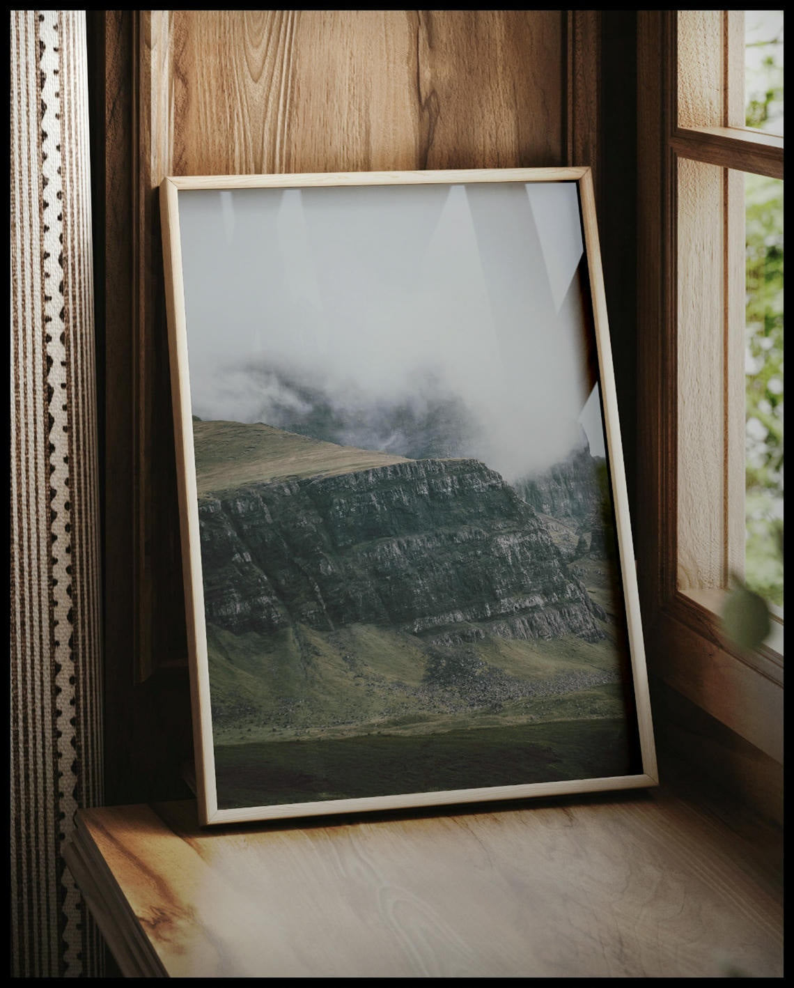 Ein gerahmtes Poster von einem steilen Abhang eines Berges, der von Gras bedeckt und von Nebel umschlungen ist, vor einem Fenster an die Wand angelehnt