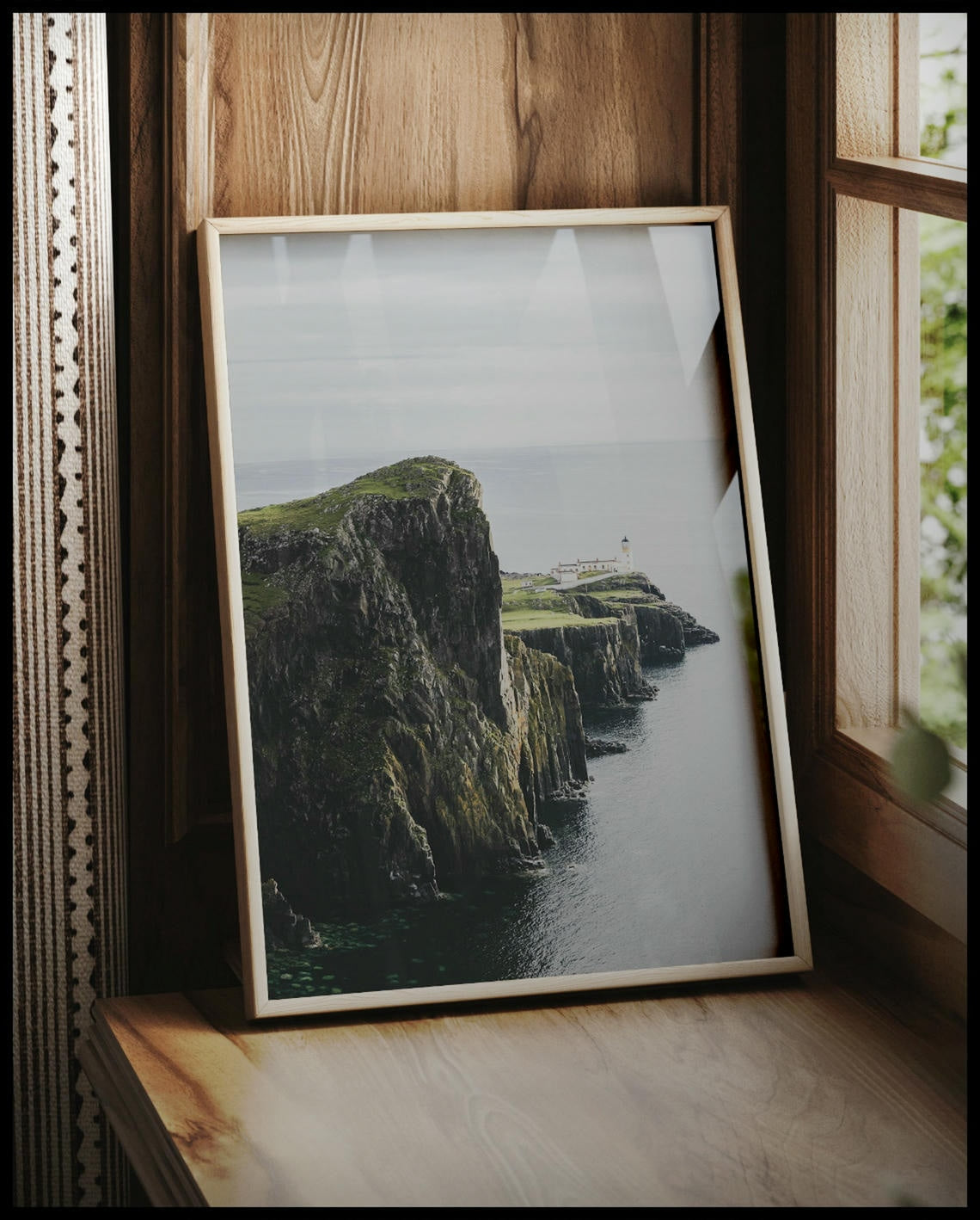 Ein gerahmtes Poster von einer kargen Klippe in Schottland mit einem Leuchtturm, vor einem Fenster an die Wand angelehnt