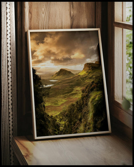 Ein gerahmtes Poster von einer kargen, grünen Berglandschaft im Sonnenuntergang, vor einem Fenster an die Wand angelehnt