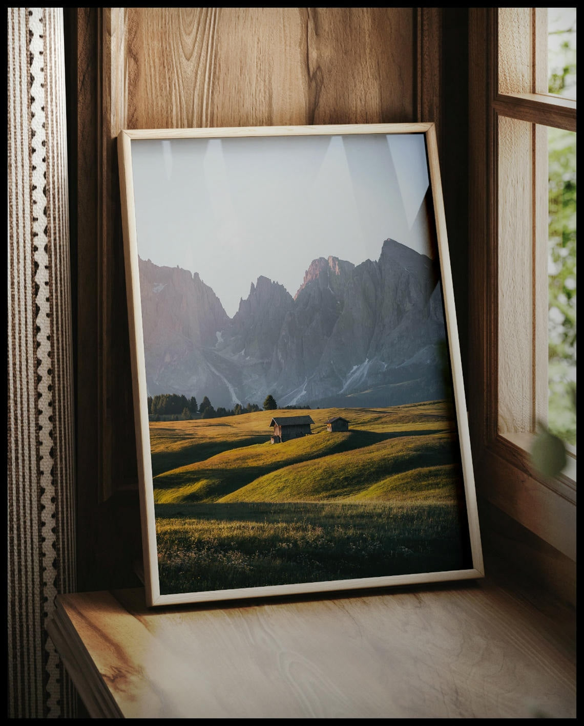 Ein gerahmtes Poster von zwei Holzhütten auf einer goldenen Wiese vor einer steilen, kargen Berglandschaft, vor einem Fenster an die Wand angelehnt