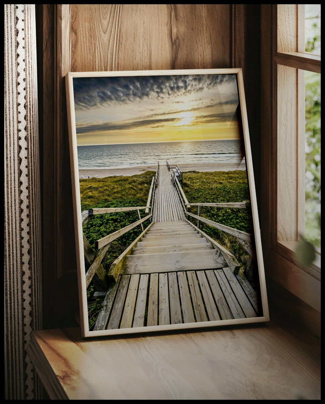 Ein gerahmtes Poster von einem Holzsteg mit einer Treppe, der durch Dünen zum Strand führt, vor einem Fenster an die Wand angelehnt