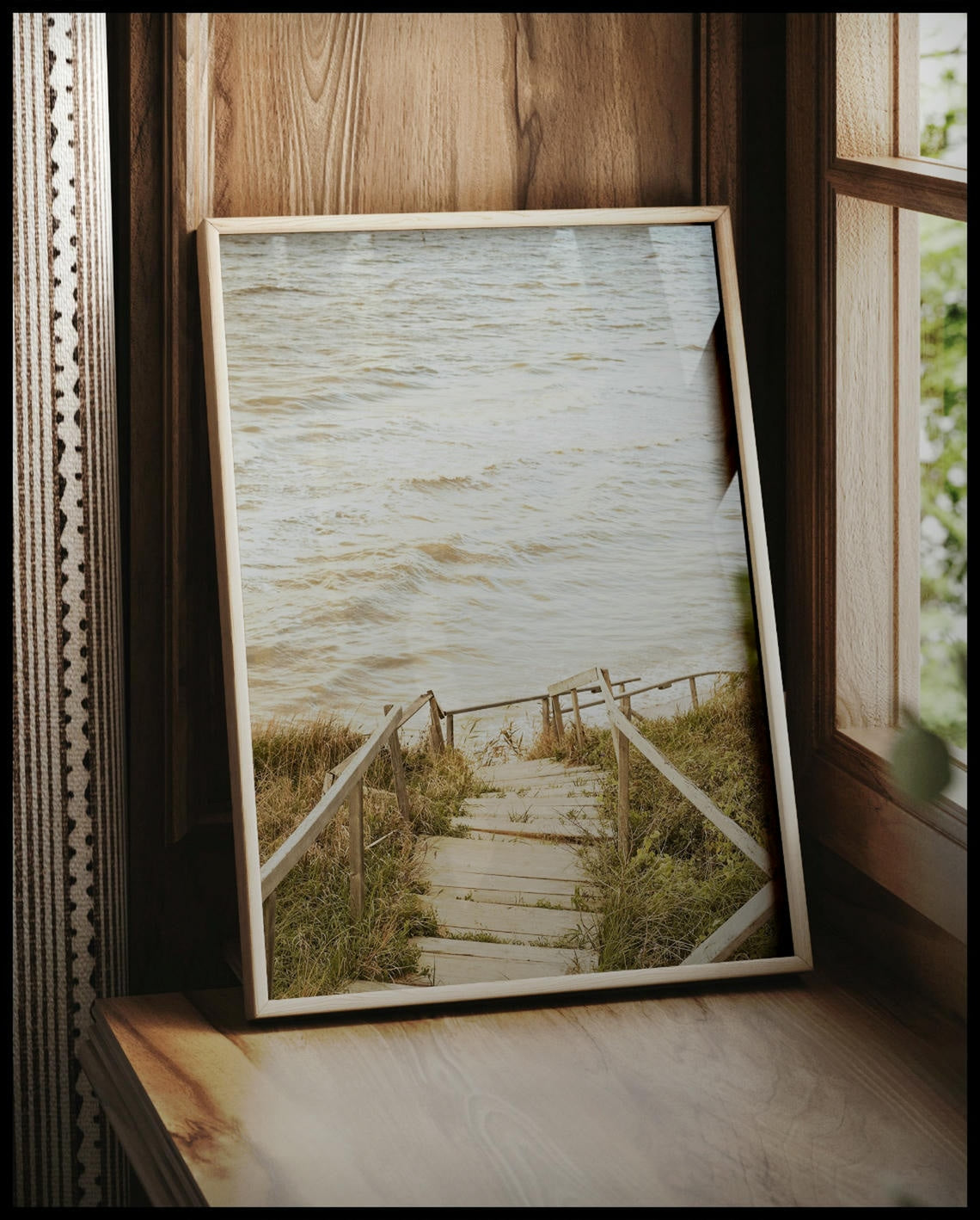 Ein gerahmtes Poster von einer Holztreppe die durch Dünen runter zum Strand führt, vor einem Fenster an die Wand angelehnt