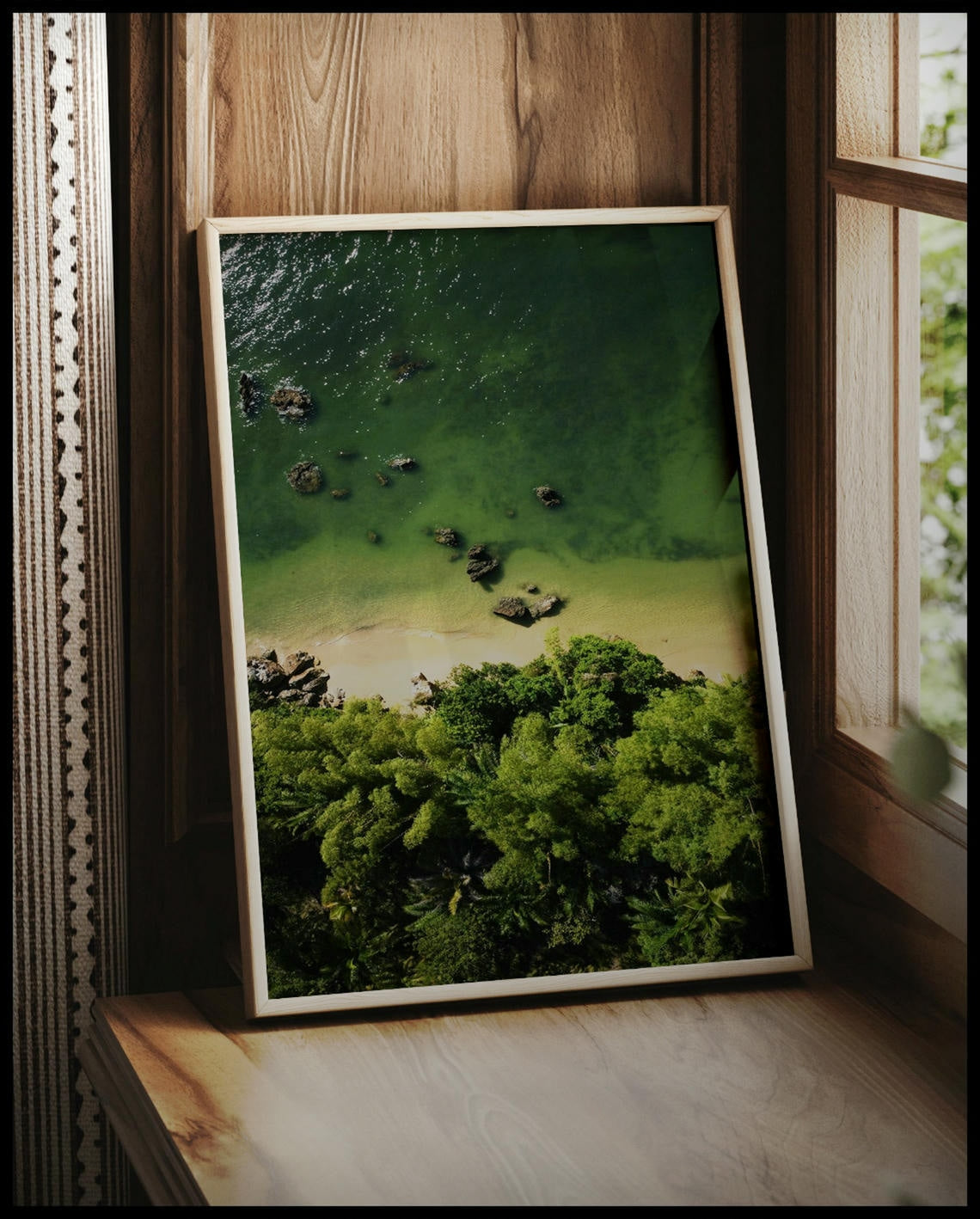 Ein gerahmtes Poster von einem Sandstrand mit grünlichem Wasser vor einem tropischen Wald, vor einem Fenster an die Wand angelehnt