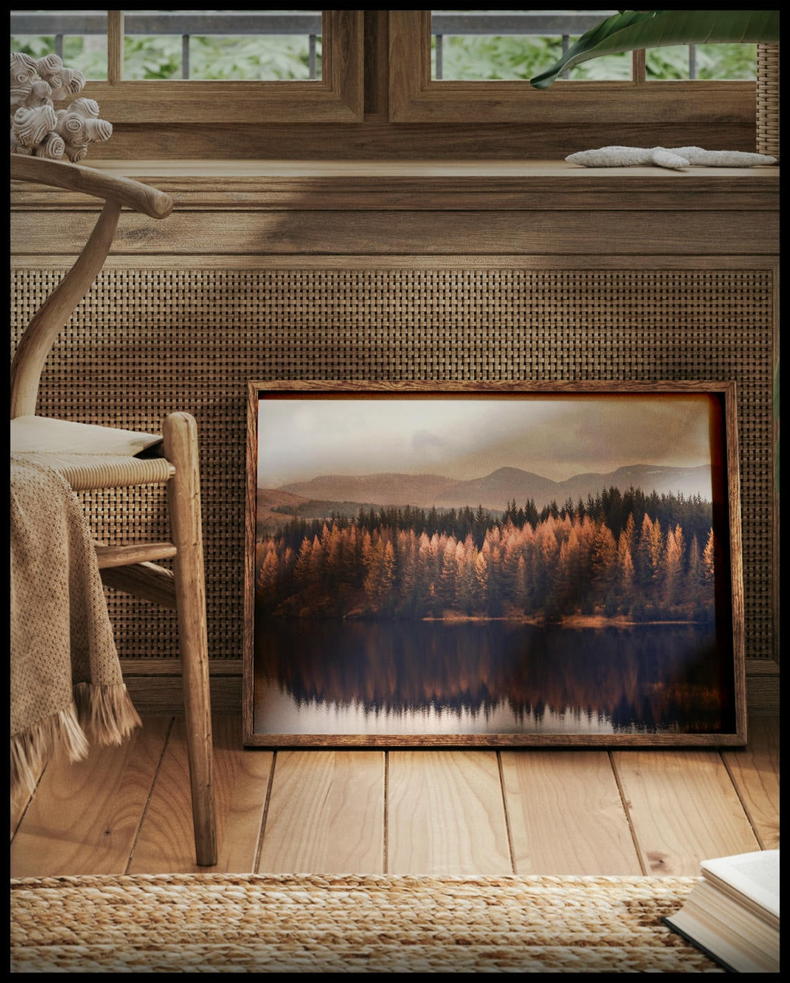 Ein gerahmtes Poster von einem herbstlichen Nadelwald, der sich in einem ruhigen See spiegelt, vor einem Fenster an die Wand angelehnt