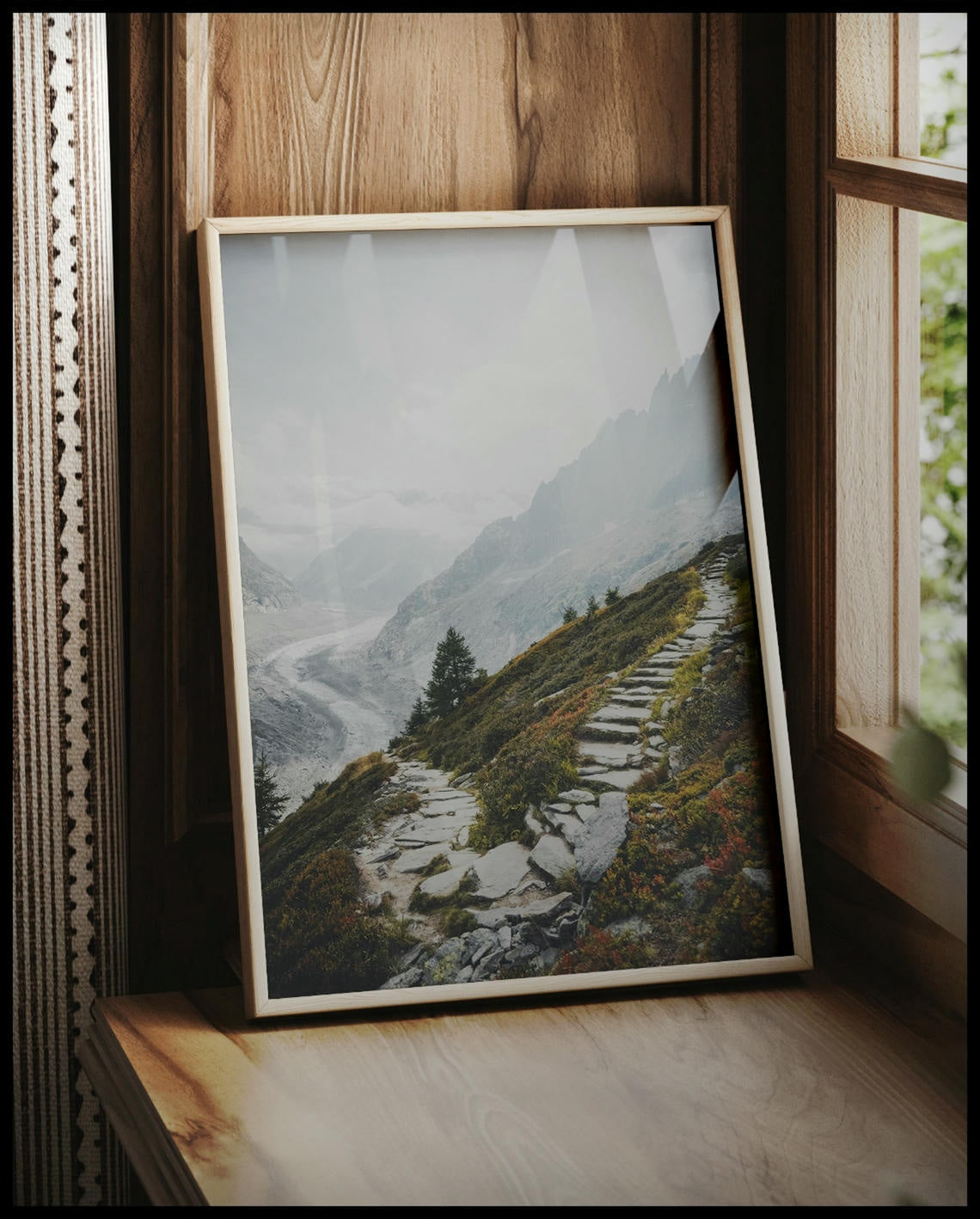 Ein gerahmtes Poster von einem Wanderweg aus steinernen Treppen inmitten einer Berglandschaft, vor einem Fenster an die Wand angelehnt