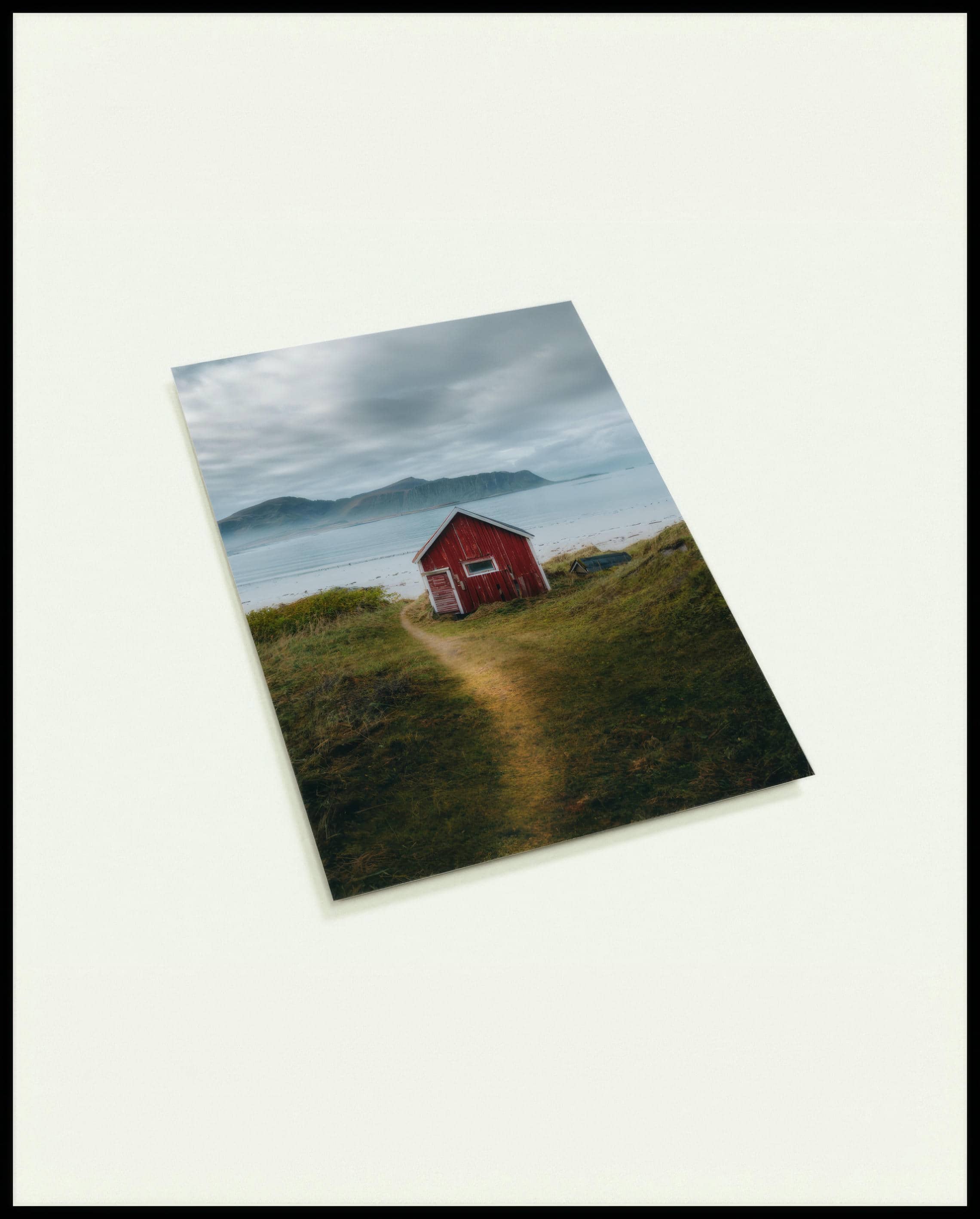 Eine Postkarte von einem kleinen roten Haus am Seeufer mit Bergen im Hintergrund.