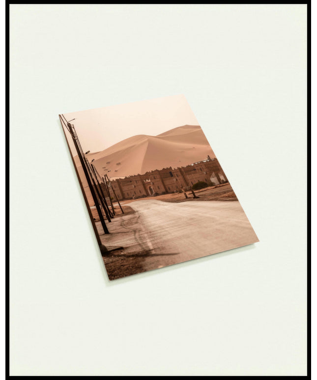 Eine Postkarte von einer Straße, die zu einer traditionellen Siedlung am Rande einer weitläufigen Wüstenlandschaft mit hohen Sanddünen führt.