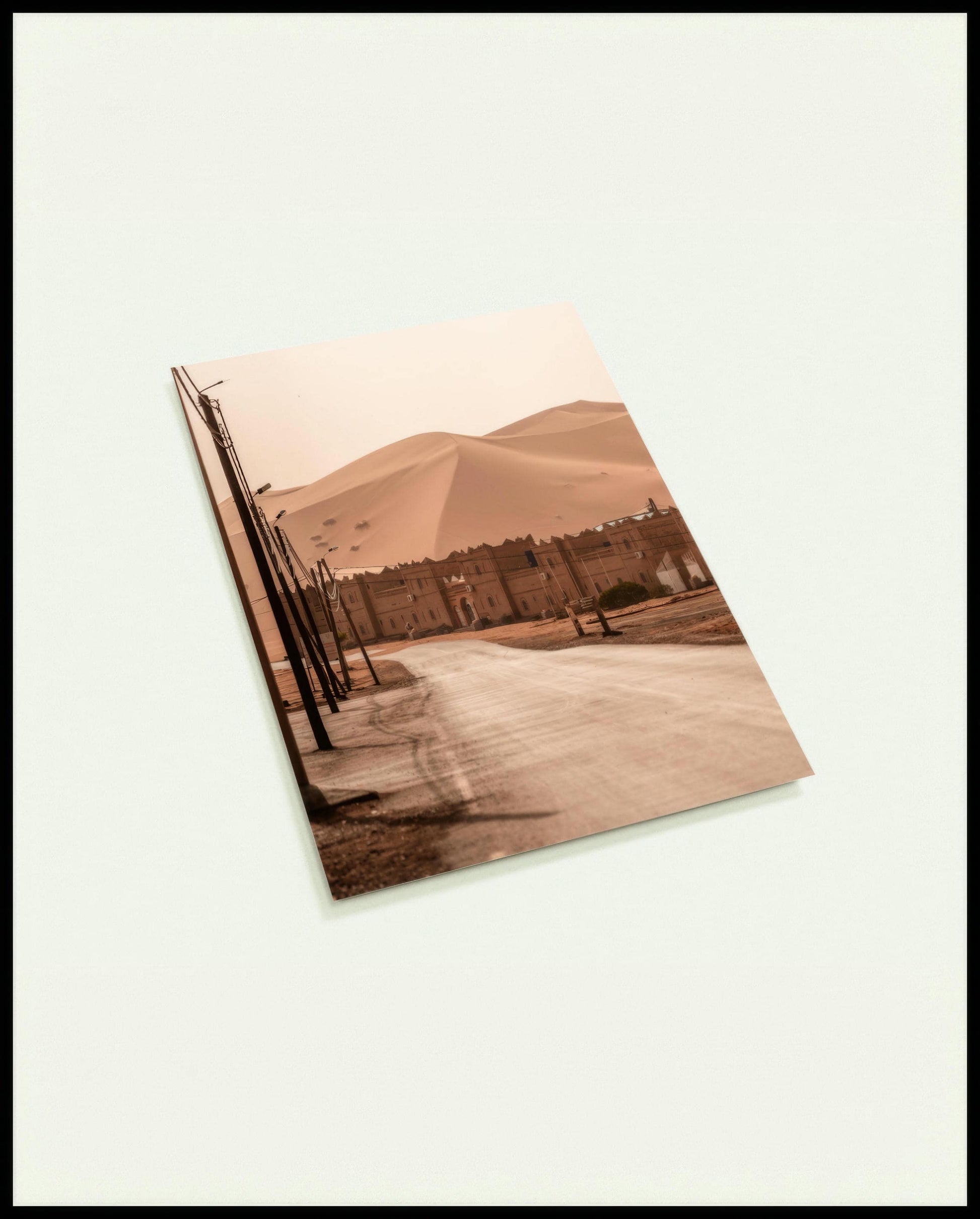 Eine Postkarte von einer Straße, die zu einer traditionellen Siedlung am Rande einer weitläufigen Wüstenlandschaft mit hohen Sanddünen führt.