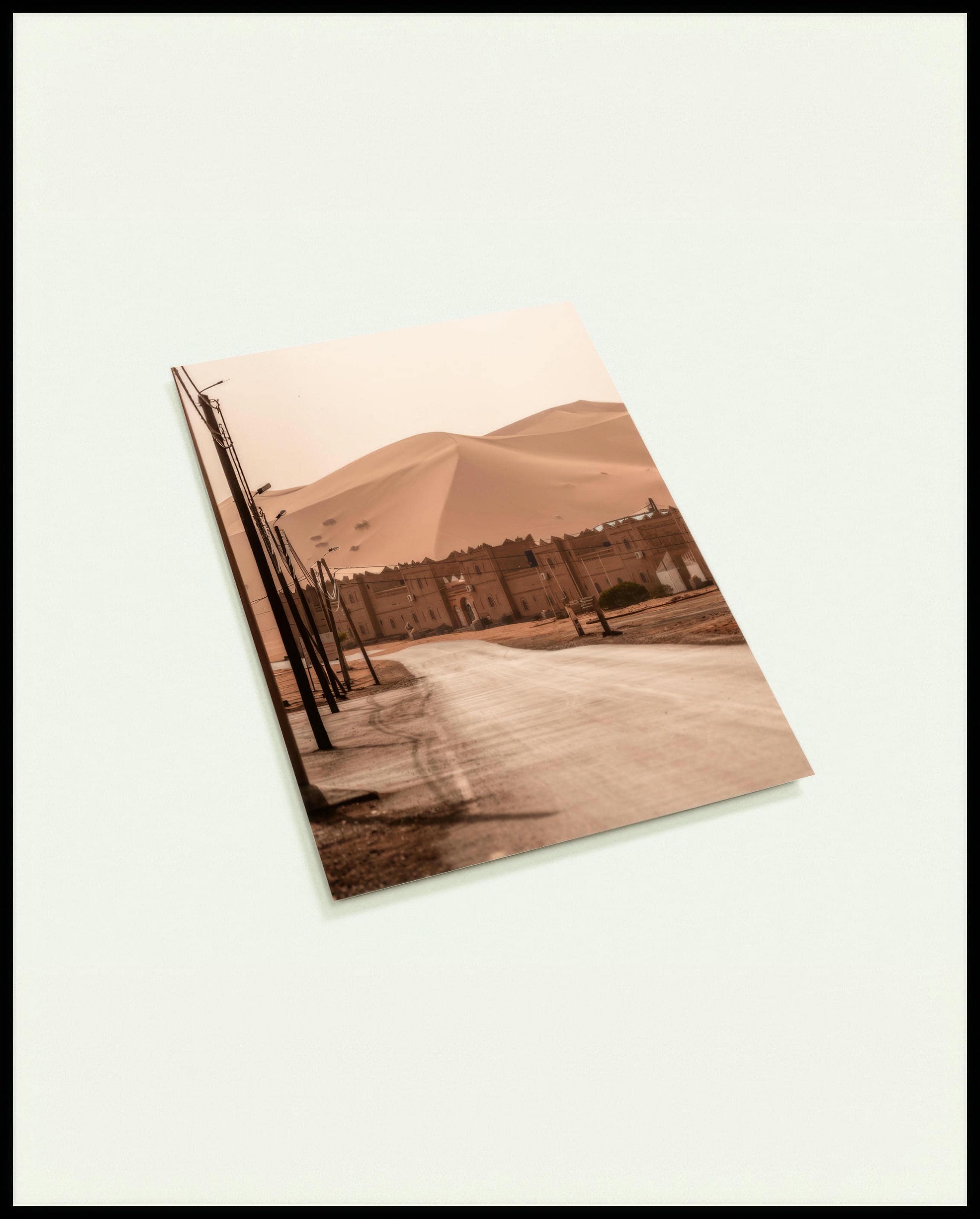 Eine Postkarte von einer Straße, die zu einer traditionellen Siedlung am Rande einer weitläufigen Wüstenlandschaft mit hohen Sanddünen führt.