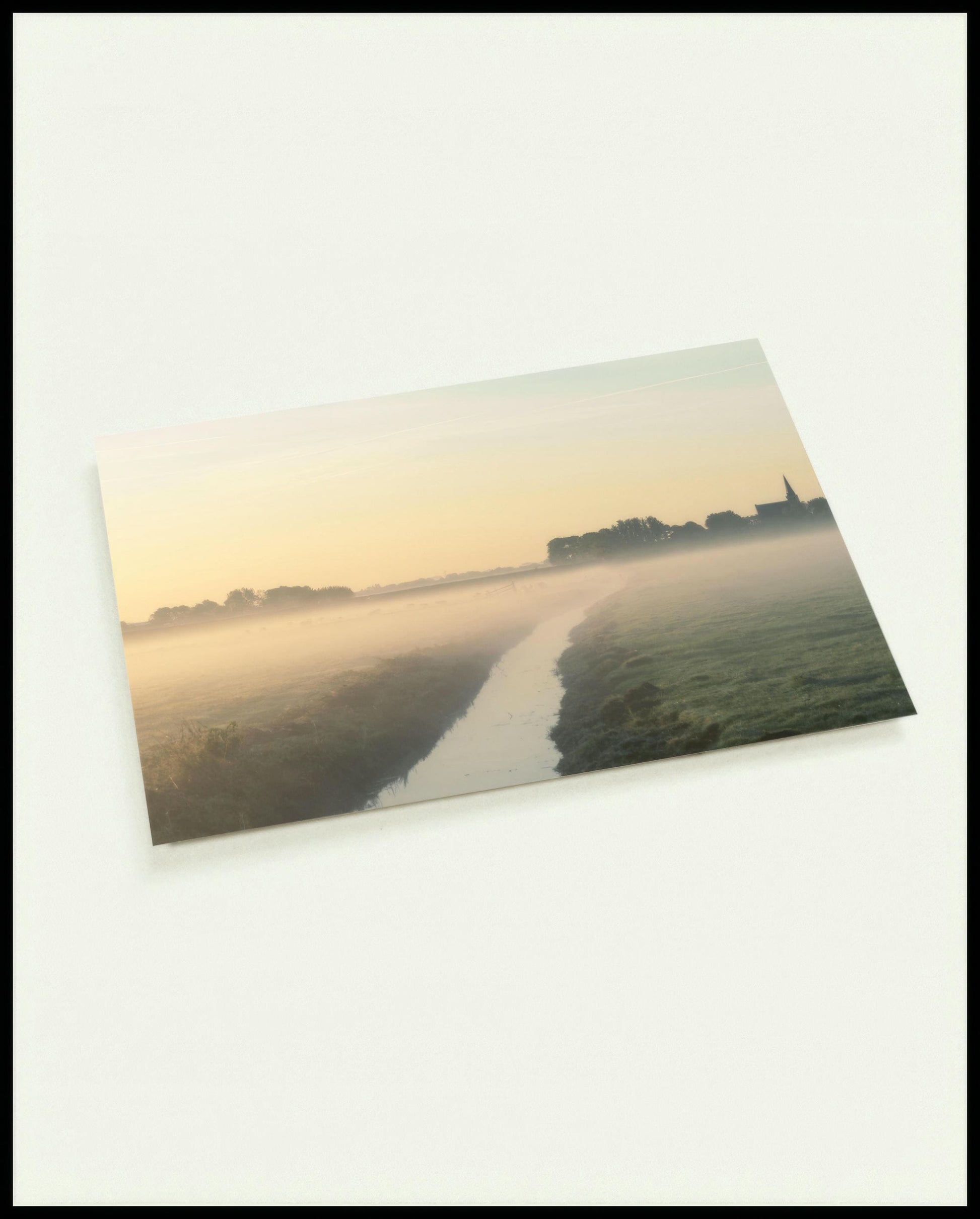 Eine Postkarte von einer nebligen Landschaft in den Niederlanden, mit einem Wasserkanal und einer Kirche im Hintergrund.