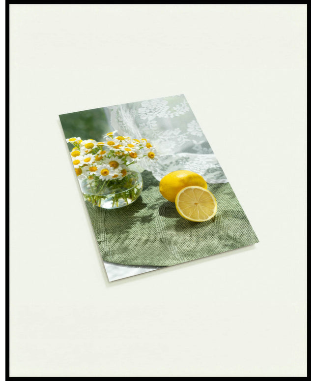 Eine Postkarte von zwei Zitronen und Blumen in einem Glas auf einer grünen Unterlage aus Stoff.