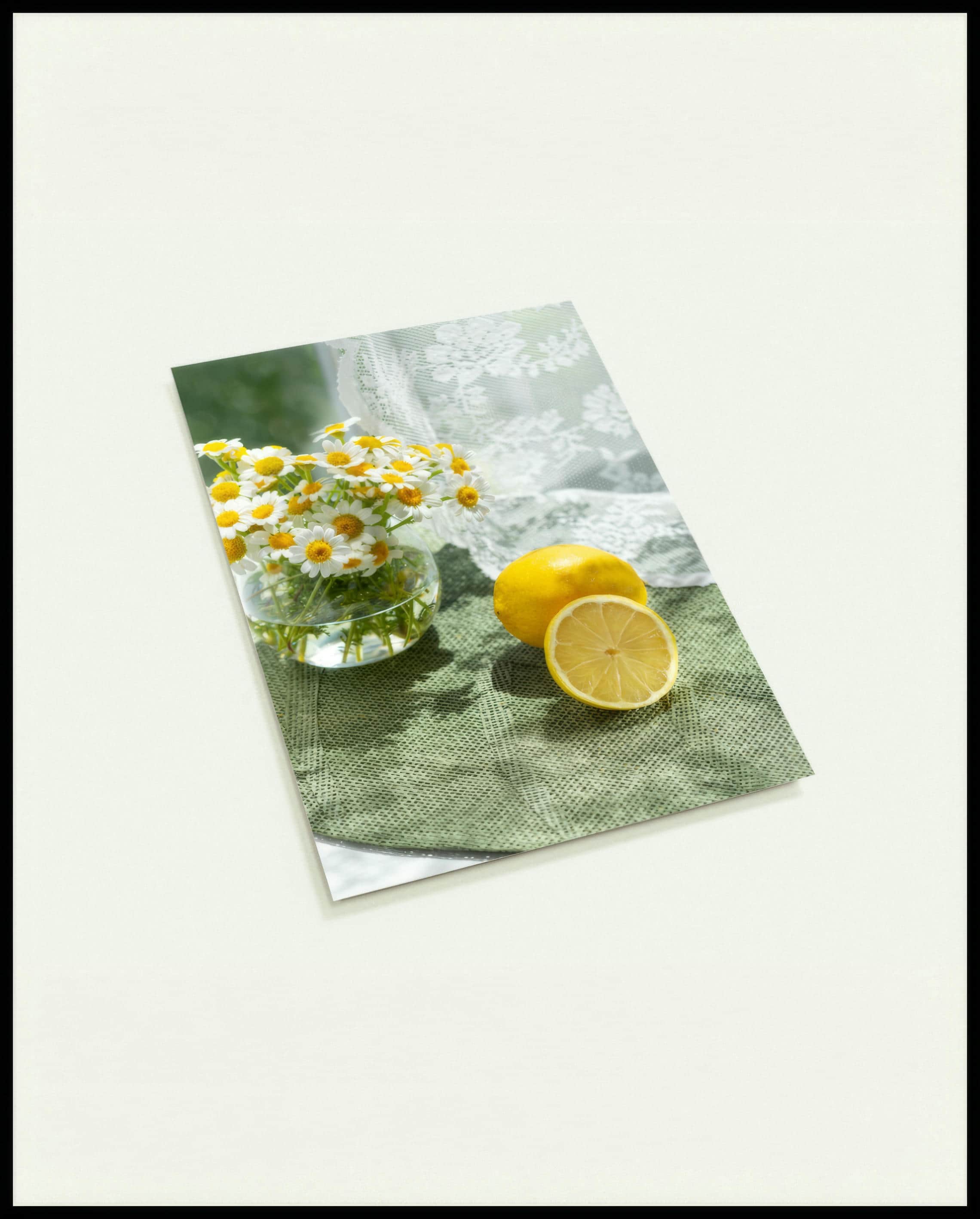 Eine Postkarte von zwei Zitronen und Blumen in einem Glas auf einer grünen Unterlage aus Stoff.
